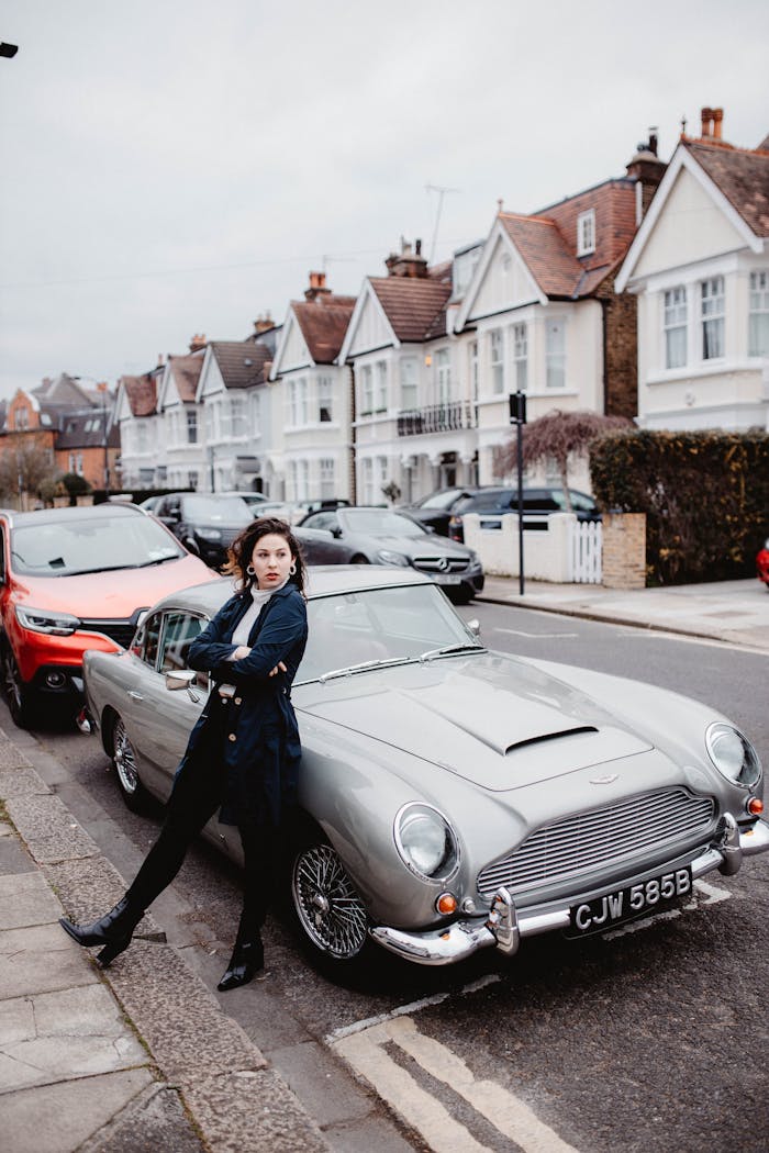 A fashionable woman stands next to a vintage car on a charming suburban street.
